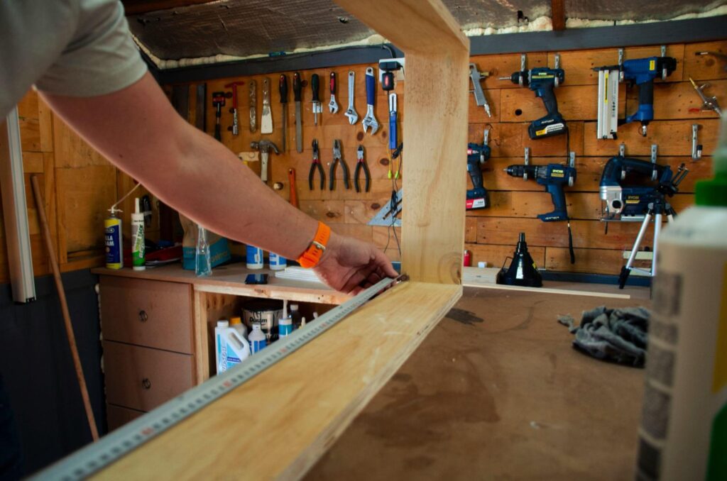 A carpenter measuring a piece of wood in a workshop. Other carpentry tools are visible in the background.